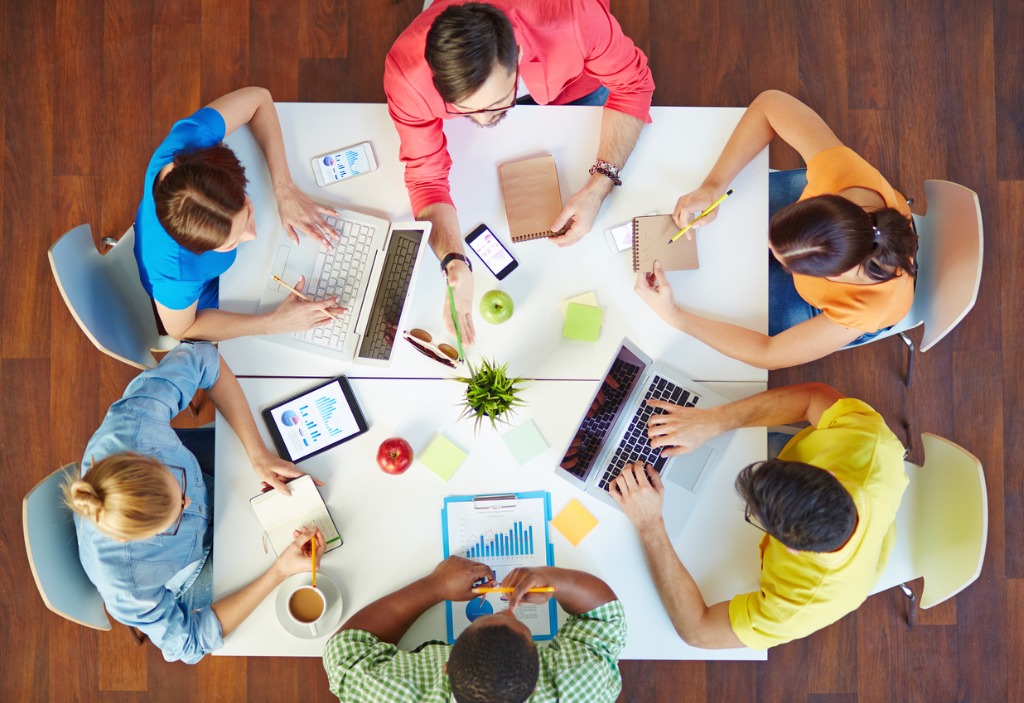 A birds-eye view of six adults gathered around a square table working and discussing. Some are writing on notepads, some are typing on computers.