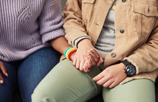 Image of two people holding hands. The person on the left is wearing a purple sweater, jeans, and a rainbow bracelet. The person on the right is wearing a brown jacket, a gray shirt, light green pants, and a black watch.