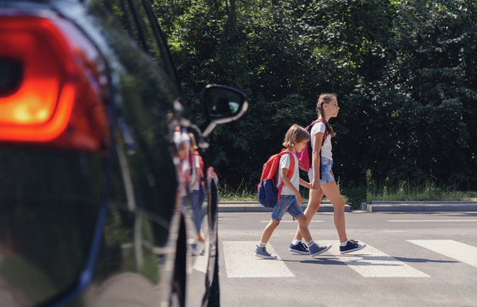 On the left, a car is stopped at a crosswalk. In the middle, two children are crossing the street on a crosswalk.