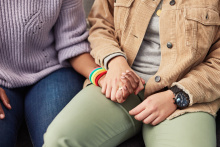 Image of two people holding hands. The person on the left is wearing a purple sweater, jeans, and a rainbow bracelet. The person on the right is wearing a brown jacket, a gray shirt, light green pants, and a black watch.